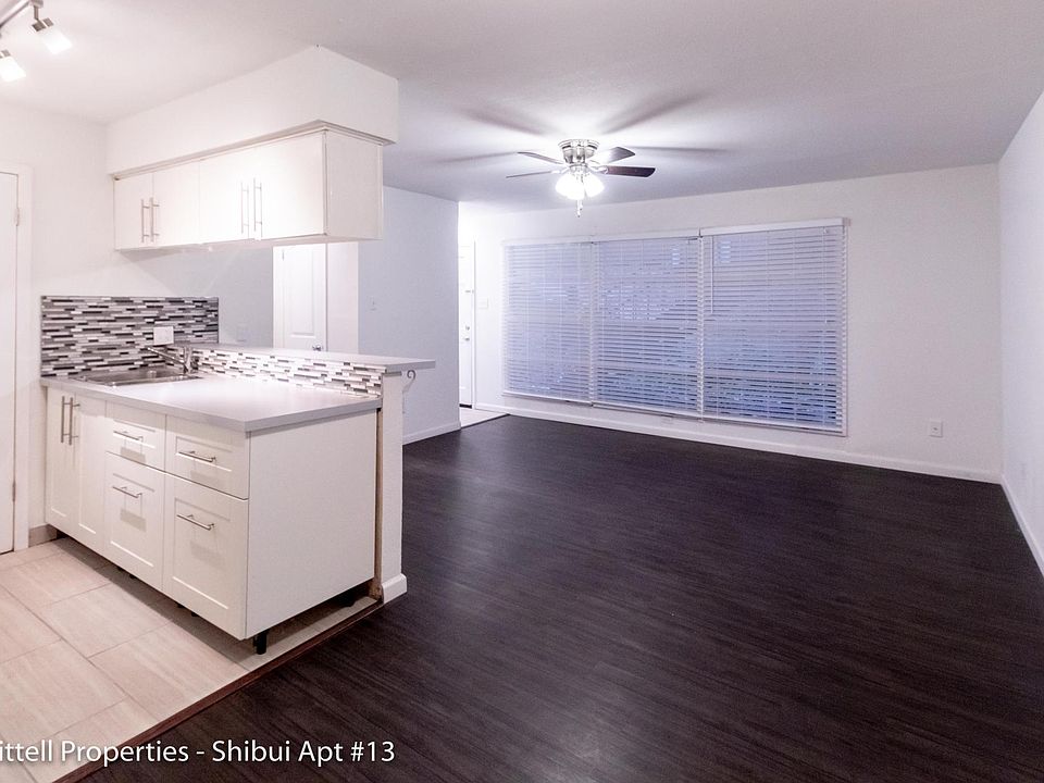 Living area, dark wood floors and large window with blinds. Ceiling fan above living area. View of kitchen space with cabinet storage above and below countertops.
