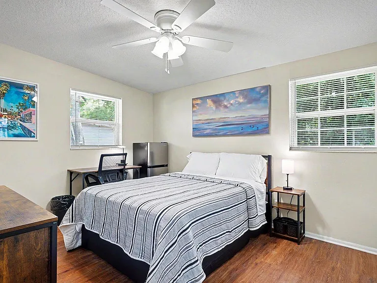 Large, furnished bedroom w/full bed, nightstand, ceiling fan, and two windows, plus a desk, dresser, and in-room mini fridge.