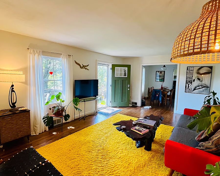 Cozy living room offers light, color, and style with a comfy sofa, burl wood coffee table, yellow shag rug, smart TV, MCM furniture, plants, and a remote speaker for your music.