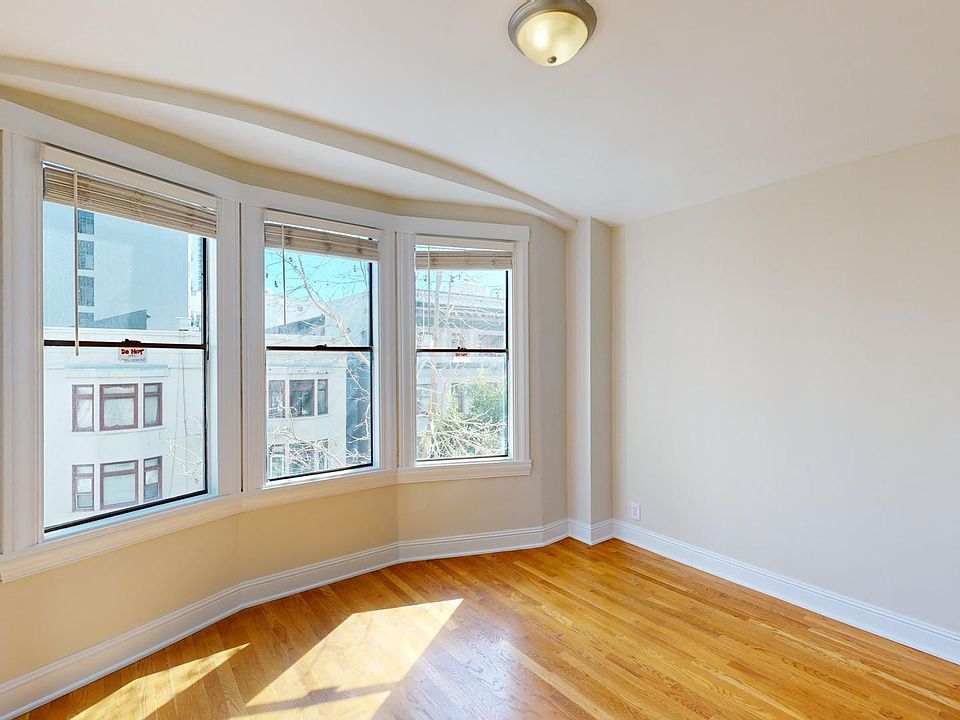 Bright, empty room with warm wood floors and cream walls. Three large bay windows let in sunlight, offering a view of neighboring buildings. Cozy and inviting space.