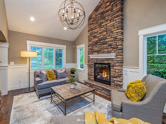 Formal living room with vaulted ceiling is anchored by floor to ceiling stacked stone gas fireplace, buffered by wainscotting trim and plantation shutters