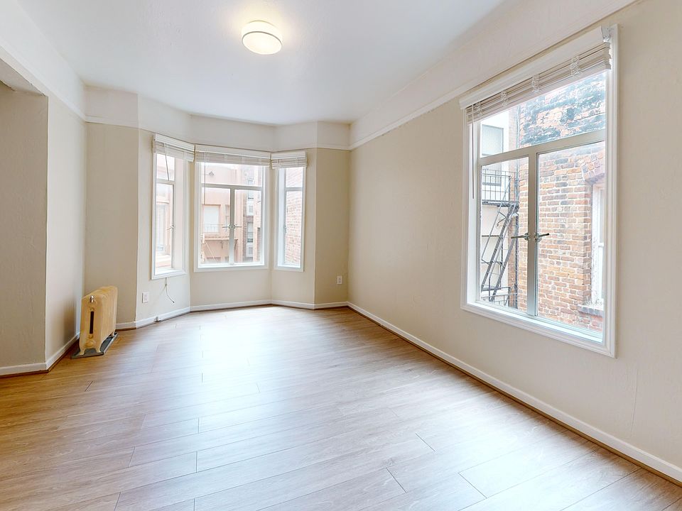 Bright, empty room with hardwood floors and large windows showcasing a view of a brick wall and fire escape.