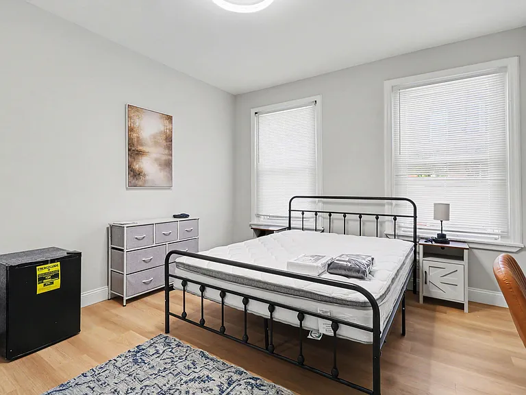 Furnished bedroom with a full bed, nightstand, dresser, and two windows with blinds for privacy and daylight.