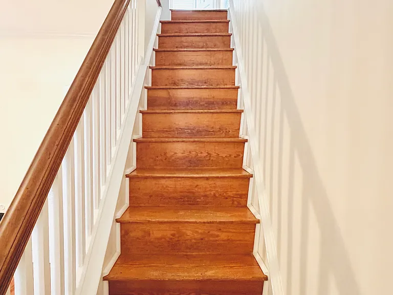 Second-floor hallway with original hardwood floors, wall sconce, decorative trim, and radiator leading to a sun-lit bedroom.