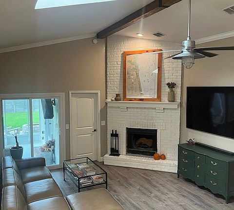 Bright and inviting open-concept living space featuring vaulted ceilings with exposed beam detail, a white brick fireplace, and abundant natural light. A glass door leads directly to the screened-in patio, creating a seamless indoor_outdoor flow.