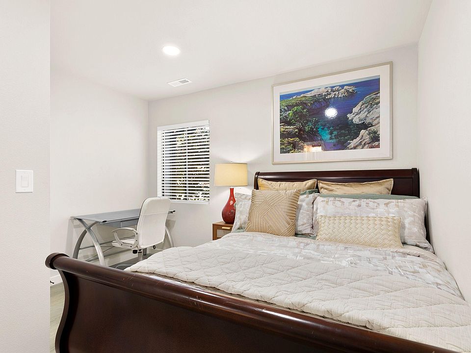 Furnished bedroom with a queen bed, nightstand lighting, and a compact desk area next to a window with blinds.