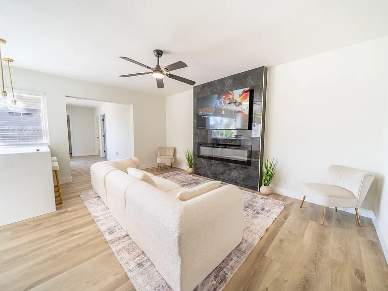 Open-concept living area with ceiling fan, large window, and media wall with fireplace, plus space for seating.