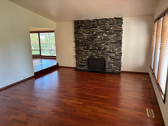 Living room from front entry. It has a vaulted ceiling, hardwood floors, and a wood burning fireplace.