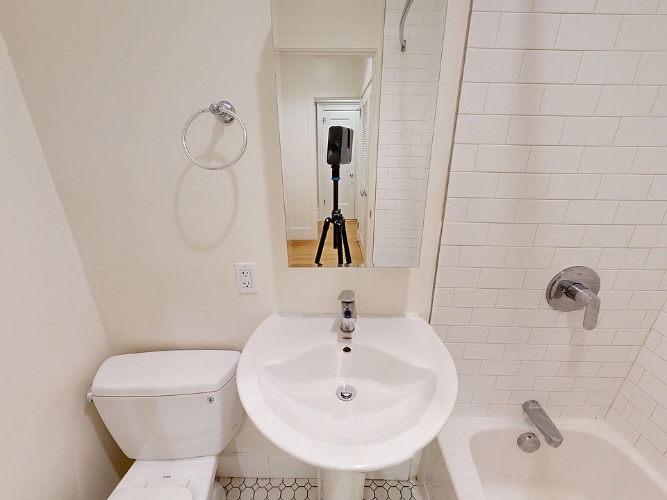 Bathroom featuring a sink, toilet, and shower with a tiled wall, reflecting a tripod and hallway in the mirror