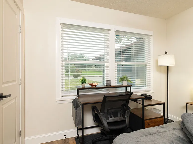 Dedicated workspace in bedroom 2 with a large desk, ergonomic chair, and natural light from double windows. Features plants, storage, and warm lamps_perfect for remote work or study.