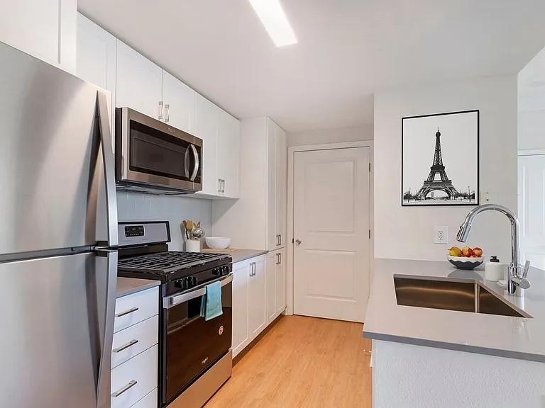 Finish Package II kitchen with white cabinetry, grey quartz countertops, extended hard surface flooring in select units, stainless steel appliances, and tile backsplash