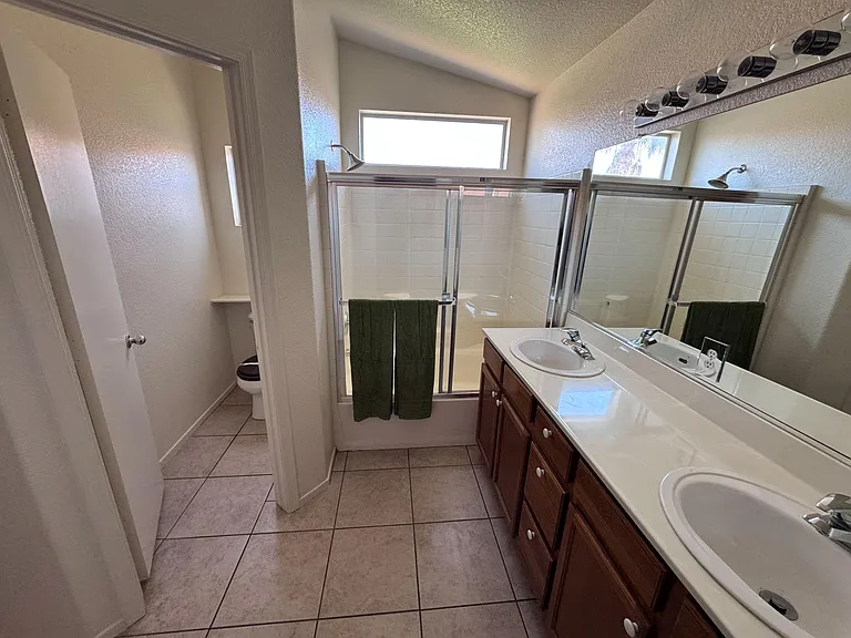 Full bathroom with dual-sink vanity, large mirror, and a tiled tub/shower with sliding glass doors and linen shelving.