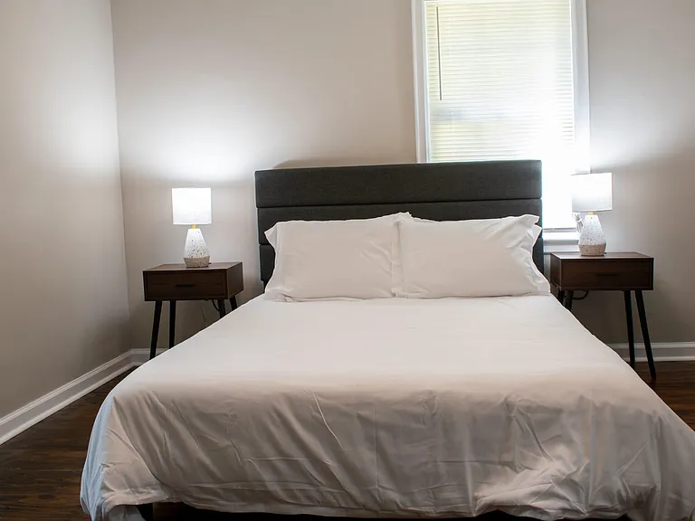 Furnished bedroom with a queen bed, upholstered headboard, matching nightstands, and bedside lamps near a window.