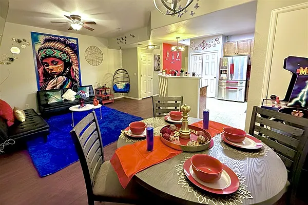 This shared area is vibrant and eclectic with bold decor, including a colorful mural & hanging chair. The dining area has a wooden table set with red dishware, candles, & a gold centerpiece.