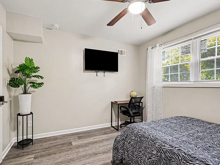 Bedroom features a ceiling fan, wall-mounted TV, and a work desk by the window with blinds for light control.