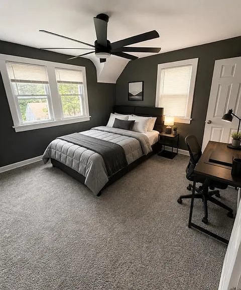 Spacious bedroom with carpet flooring, ceiling fan, dual windows, and a dedicated desk area for working or studying.