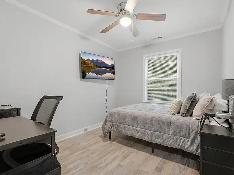 Well-appointed bedroom with a full bed, work desk, wall-mounted TV, and ceiling fan plus a window with blinds.