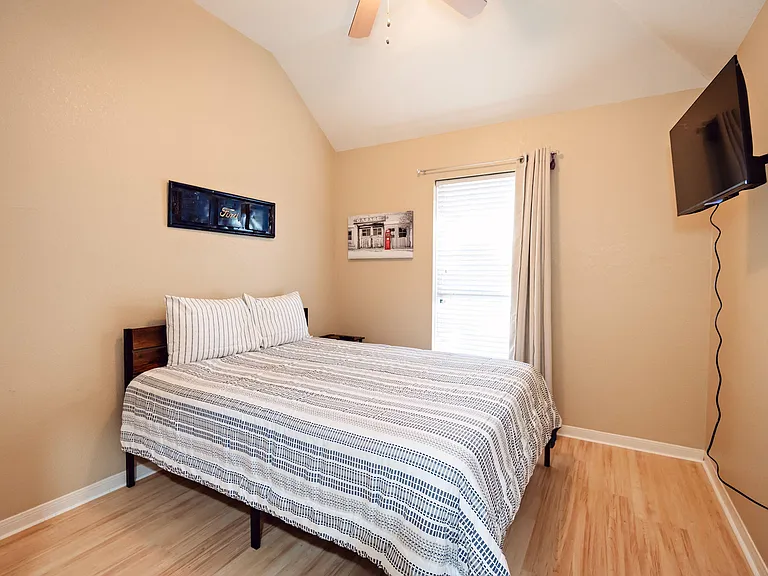Furnished bedroom with a full bed, wood-style flooring, window with blinds and curtains, and a wall-mounted TV.