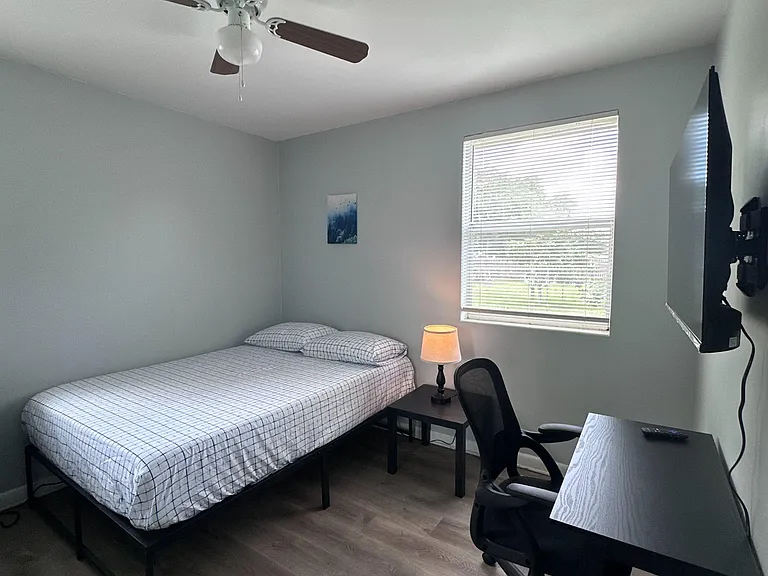 Furnished bedroom with ceiling fan, large window with blinds, work desk, and wall-mounted TV on durable flooring.