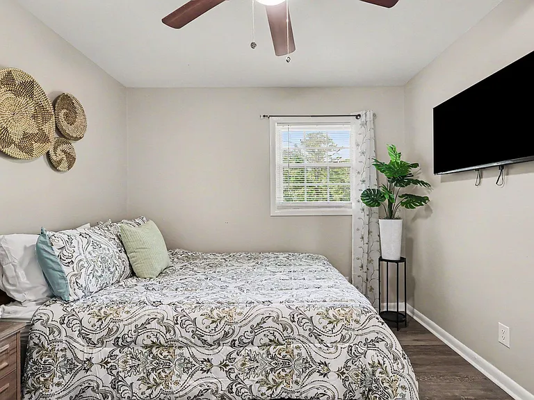 Furnished bedroom with a comfortable bed, ceiling fan, window with blinds, and wall-mounted TV for easy relaxing.