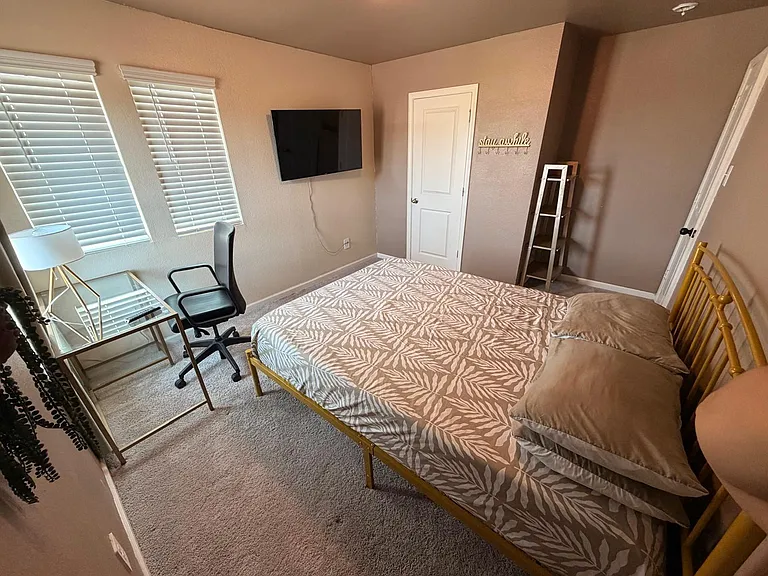 Furnished bedroom with queen bed, work desk, wall-mounted TV, and dual windows with blinds for natural light.