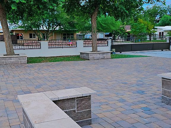 Expansive paver courtyard with mature trees and custom seating planters, offering shade and a serene view toward the gated front entrance.