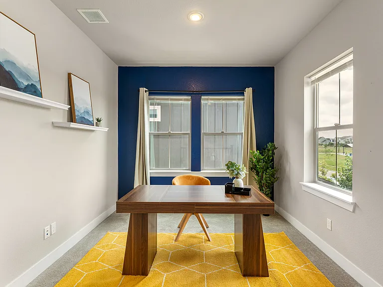 A cozy and modern home office featuring a natural wooden desk and a comfortable ergonomic chair. The space is energized by a vibrant mustard-yellow rug that adds warmth and personality. A bold navy accent wall stands out, complemented by two large windows