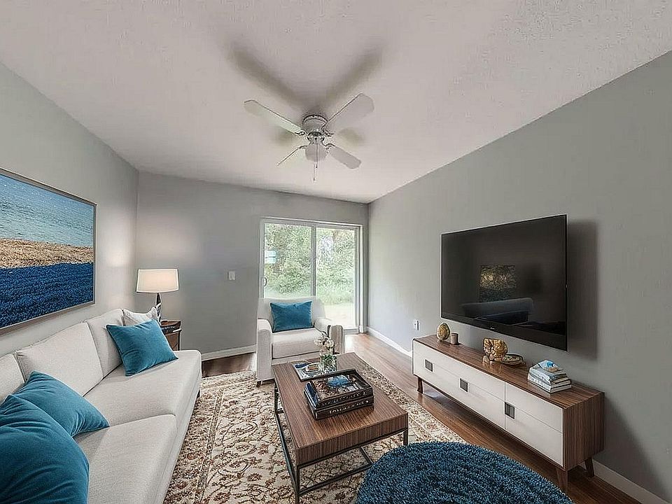 Inviting living room with modern decor, plush sofas adorned with teal cushions, elegant wood furniture, and a large flat-screen TV, offering a serene view through sliding glass doors.