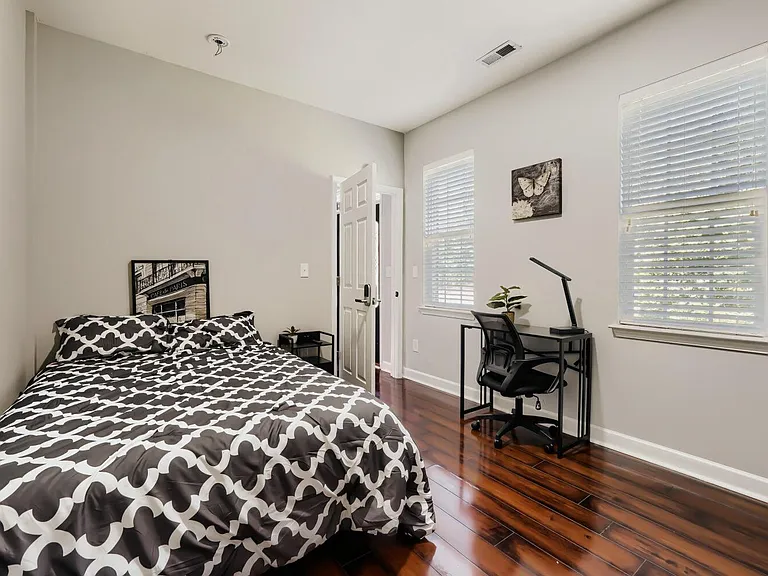 Furnished bedroom with hardwood floors, a comfortable bed, and a dedicated desk workspace beside large windows.
