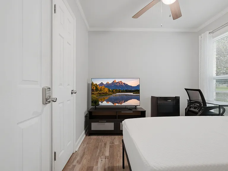 Private bedroom with a bed, TV on a media console, and a work desk beside a window with blinds and curtains.