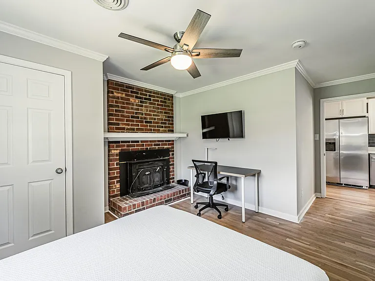 Spacious bedroom with ceiling fan, brick fireplace feature, wall-mounted TV, and a dedicated desk area near the kitchen.