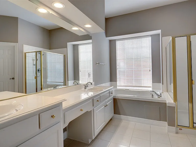 Bathroom featuring a dual vanity with integrated sinks, a built-in bathtub with a window, a glass-enclosed shower with gold-tone framing, a full-width mirror, and recessed lighting