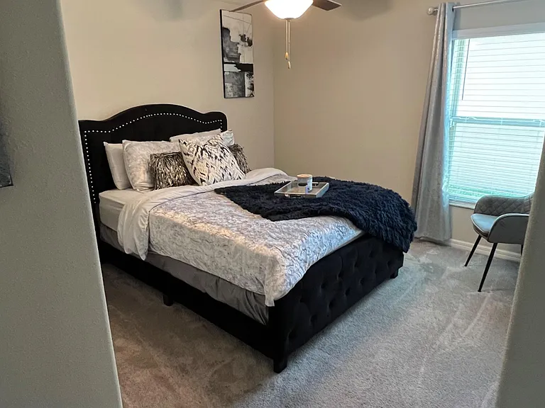 Furnished bedroom with a full bed, ceiling fan light, soft carpeting, and a window with blinds and curtains.