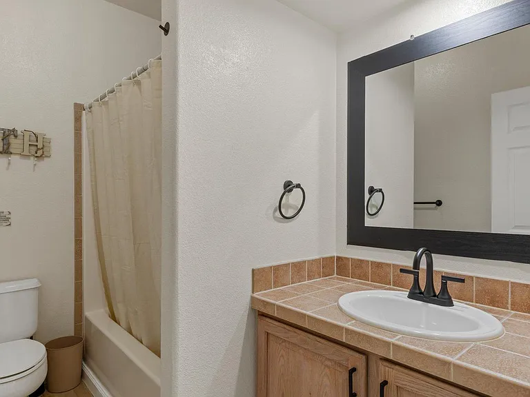 Full bathroom with tiled vanity countertop, large framed mirror, and tub/shower combo with curtain and towel storage.