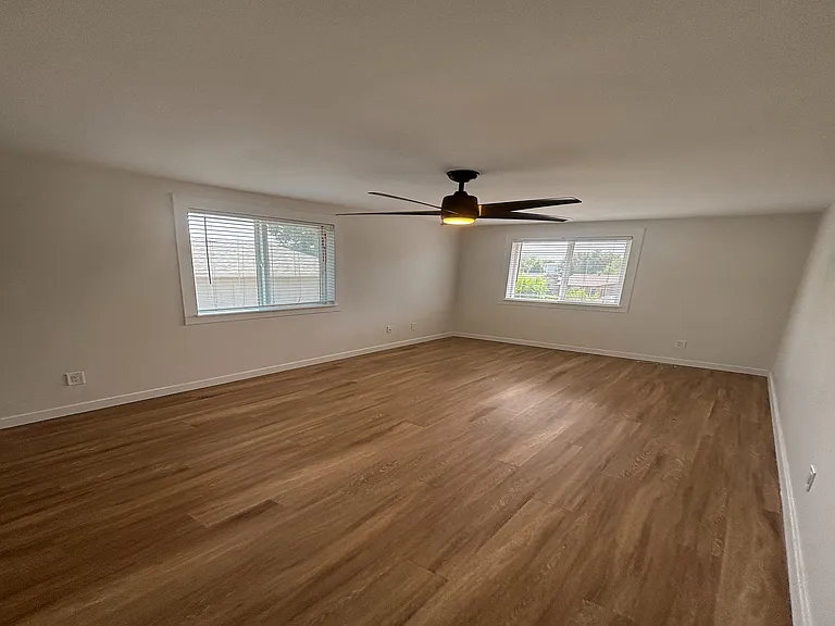 Bright and airy room with sleek wood flooring, featuring large windows and a modern ceiling fan.