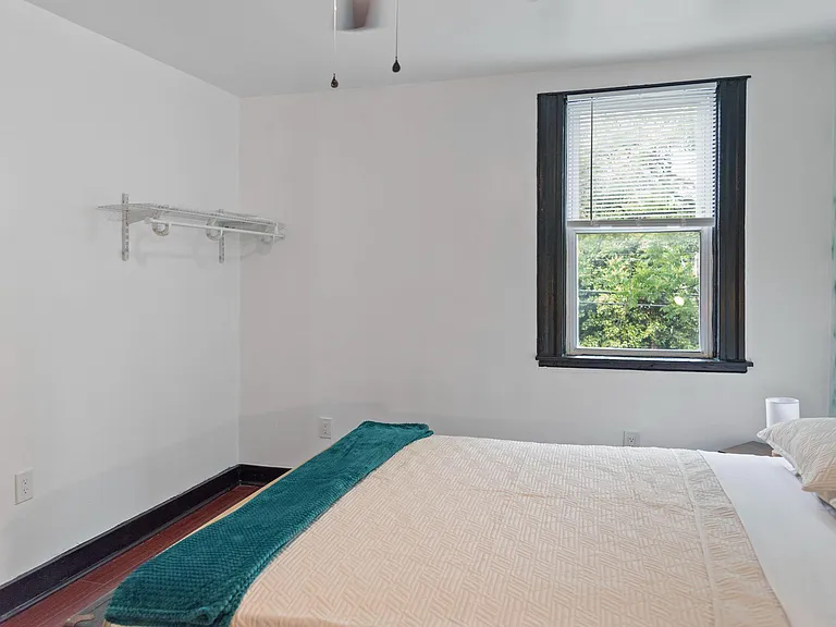 Furnished bedroom with a comfortable bed, window with blinds, and convenient wall-mounted shelving for storage.