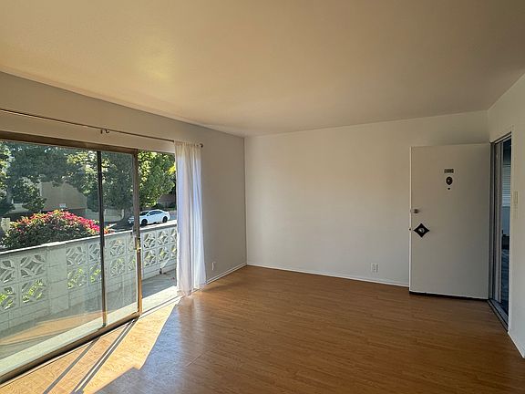 Living room with tan flooring facing the glass and metal sliding doors, with white curtains and views of the balcony.