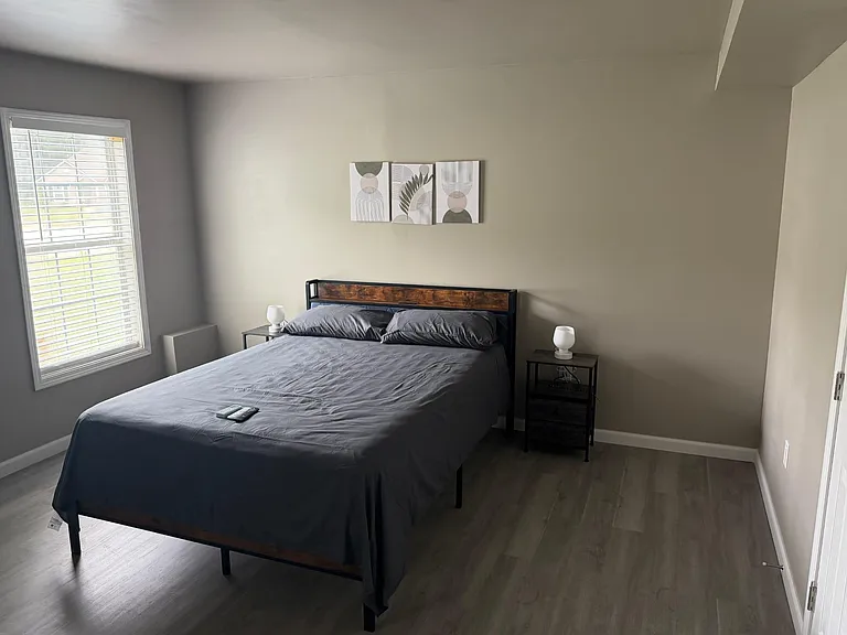 Furnished bedroom with a full-size bed, matching nightstands and lamps, and a large window with blinds for natural light.