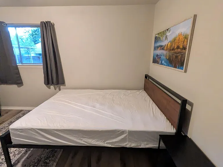 Private bedroom with a full bed, side table, and window with curtains, plus durable wood-look flooring and wall art.
