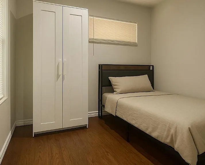 Private bedroom with a single bed, wardrobe storage, and window blinds for light control on durable wood-look flooring.