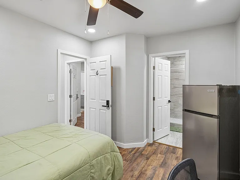 Private bedroom with wood-look flooring, ceiling fan, and convenient in-room mini fridge, plus easy access to nearby bath.