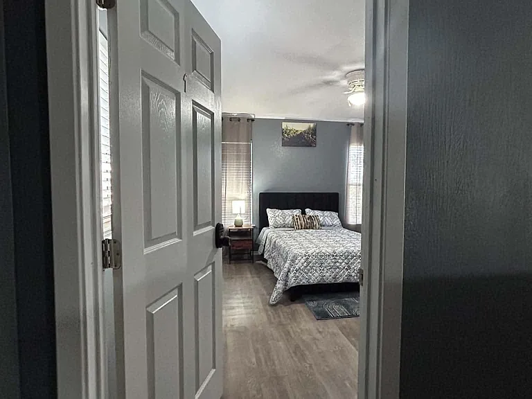 Private bedroom with wood-look flooring, a full bed, side table, and windows with blinds for natural light.