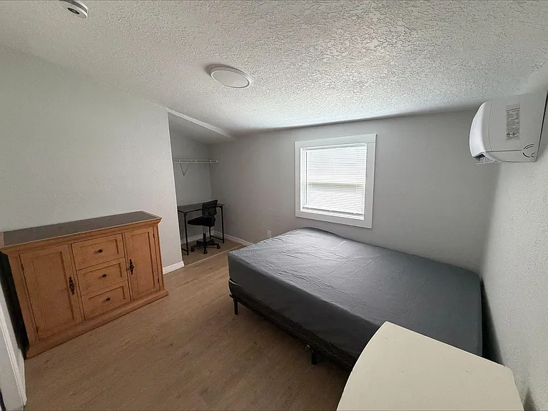 Furnished bedroom with a bed, dresser, and a dedicated desk nook plus closet shelving and a window with blinds.