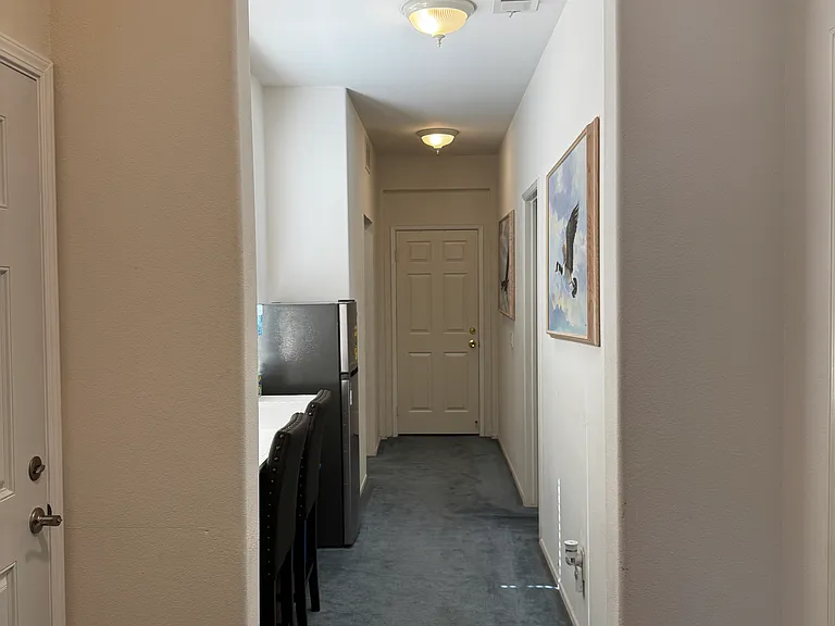 North wing hallway of home is a common area primarily used by room rental residents. Hallway consists of table-top with stools, new refrigerator, art on walls, bathroom shared by two separate rented room residents on right side by entry door.