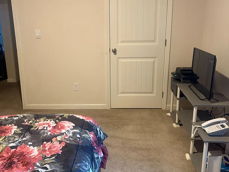 Carpeted bedroom with neutral walls, a paneled door, and space for a bed plus a small desk or media setup.