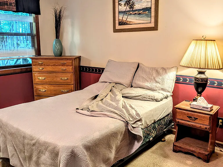 Furnished bedroom with a twin bed, nightstand lighting, dresser storage, and a window with blinds for natural light.