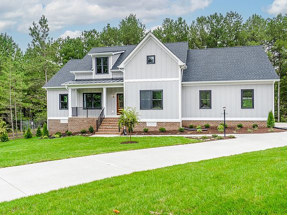 A white house with a gabled roof sits on a manicured lawn with a concrete driveway, surrounded by tr