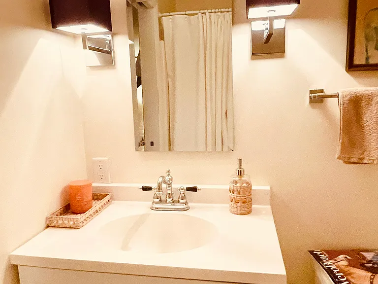 Your immaculate, dedicated private bathroom. Features a pristine white vanity, elegant traditional chrome fixtures, warm custom lighting sconces, and classic window blinds for privacy and comfort.
