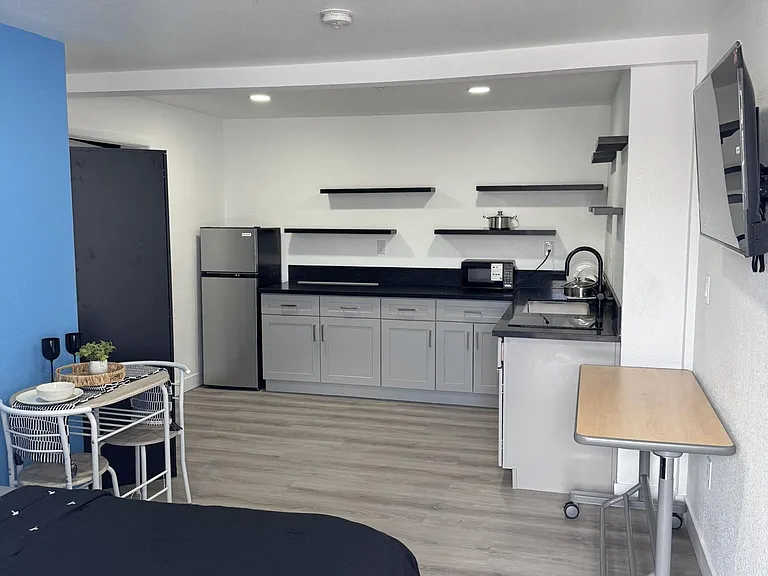 Studio-style bedroom with kitchenette, featuring ample counter space, open shelving, and a dining/work table.