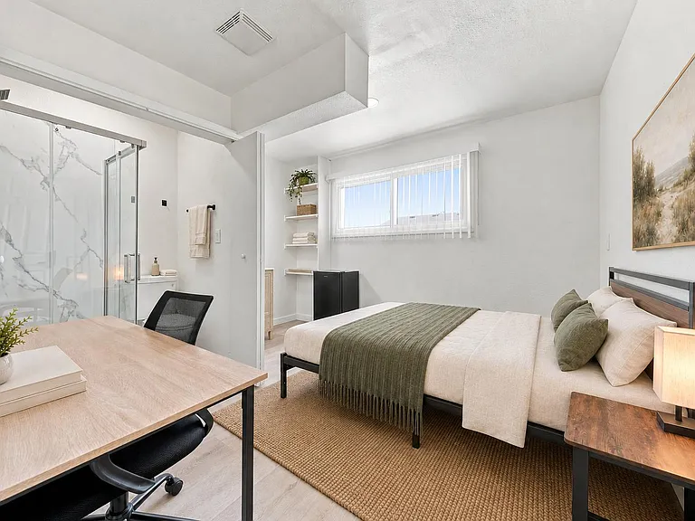Furnished bedroom with desk, mini fridge, open shelving, and an en-suite bath with glass shower and window blinds.